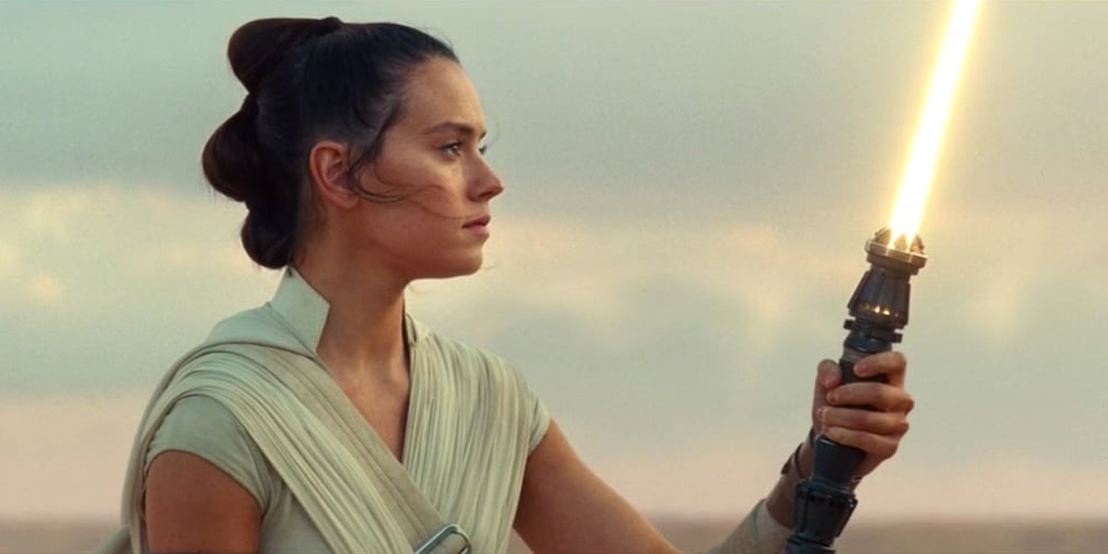 An image of Rey looking meaningfully at a yellow lightsaber she's holding in front of her.