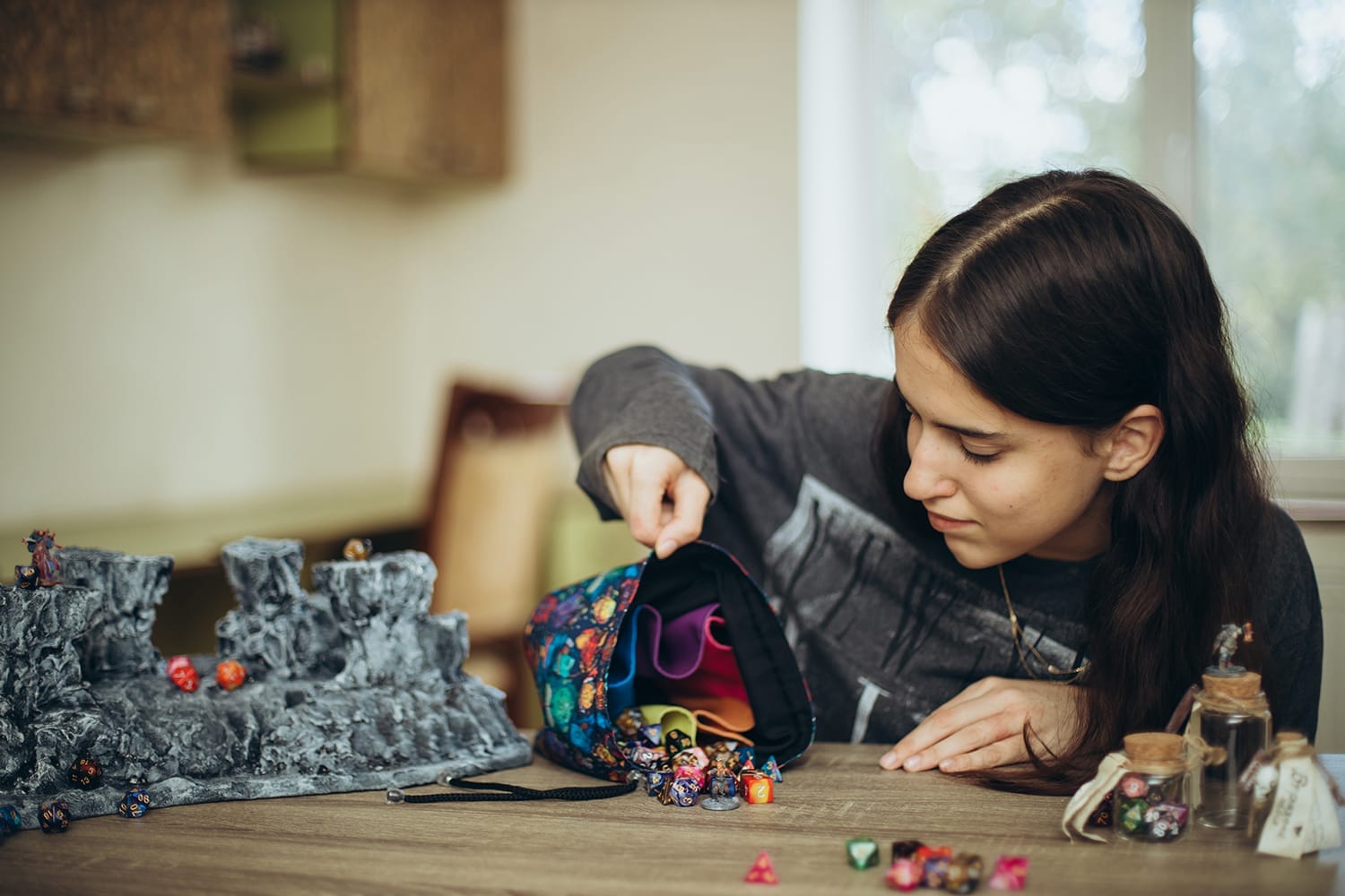 Open handmade dice bag with rainbow-colored inner pockets standing on a table, dice spilling out next to miniature terrain